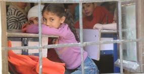 Vue sur une salle de classe. La photographie a été prise de l'extérieur, on y voit les barreaux de la fenêtre. Les enfants à l'intérieur regardent l'objectif de la caméra.