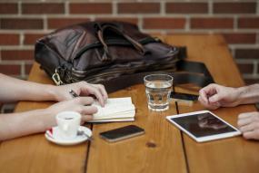 Photographie de deux personnes face à face à une table, mais on ne voit que leurs mains. Des breuvages et des appareils numériques sont posés devant elles.